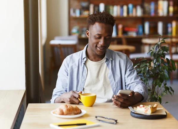 Vonzó afro-amerikai srác használ okostelefon fehér reggelizik a kávézóban — Stock Fotó