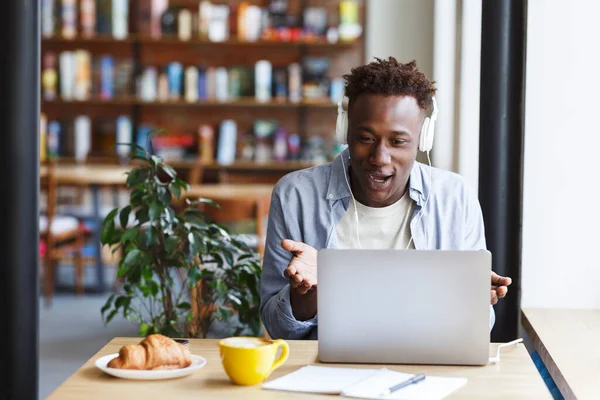Opgewonden Afro-Amerikaanse man met headset in gesprek met zijn vrienden op video chat in coffeeshop, lege ruimte — Stockfoto