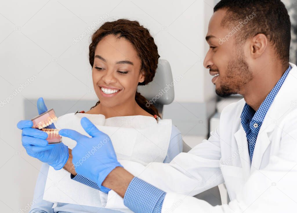 Black handsome man dentist showing female patient plastic jaw