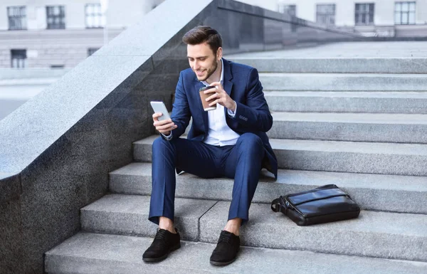 Happy businessman in formal wear drinking takeout coffee and browsing net via smartphone on stairs at lunchtime