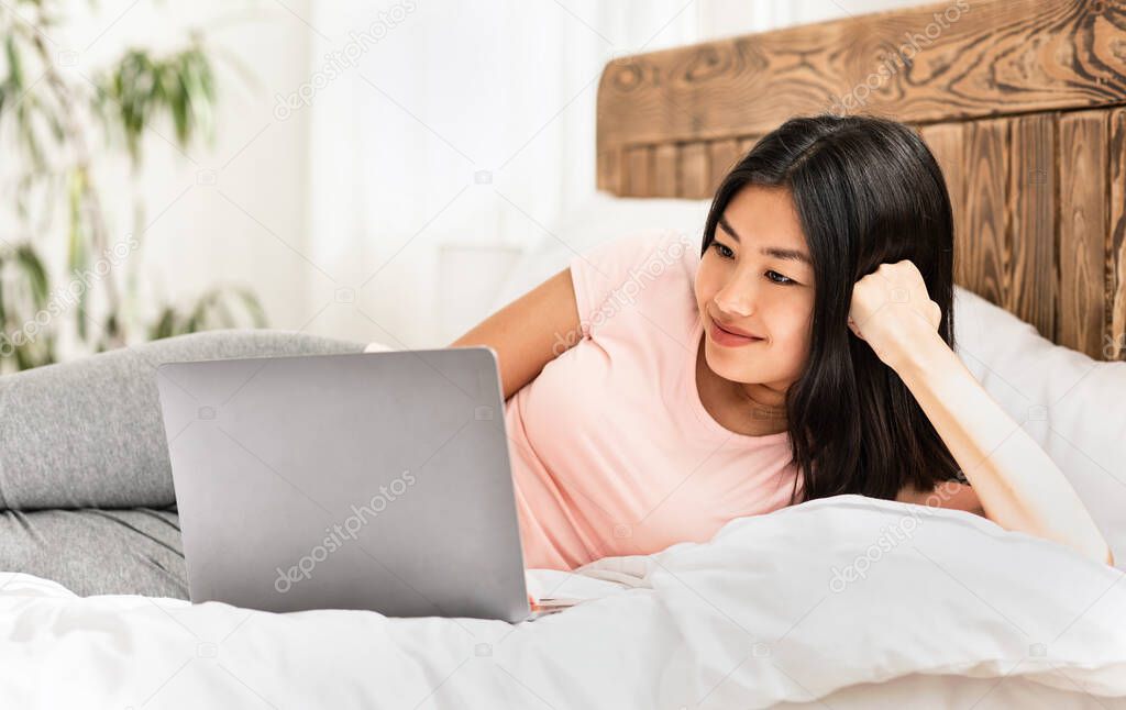 Smiling Japanese Girl Working On Laptop Lying In Bed Indoors