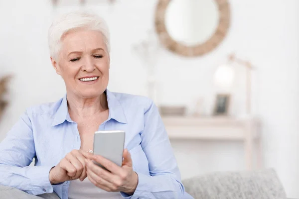 Lächelnde ältere Frau hält Handy in der Hand, liest Nachrichten, während sie zu Hause ausruht — Stockfoto
