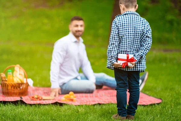 Best surprise for dad. Shy little boy with fathers day gift behind his back and parent on picnic in park