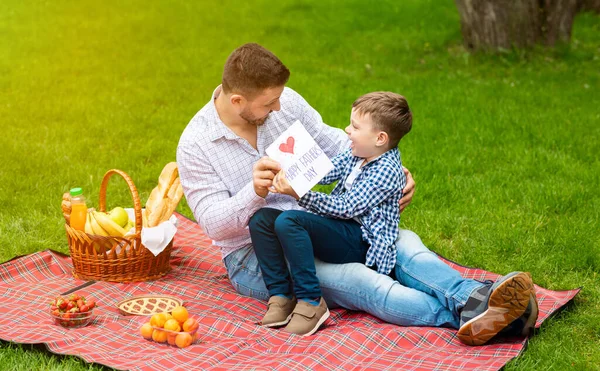 Te quiero, papi. Adorable niño dando tarjeta de felicitación al padre durante el picnic familiar en el prado verde — Foto de Stock