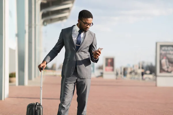 Hombre negro en viaje de negocios utilizando el teléfono móvil de pie cerca del aeropuerto — Foto de Stock