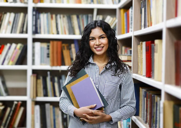 Sorridente ragazza latina in possesso di libri di testo in biblioteca — Foto Stock