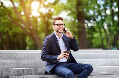 Şehir parkındaki taş merdivenlerde cep telefonuyla konuşan kahveli neşeli girişimci.