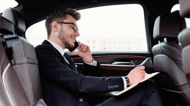 İş adamı, lüks bir arabada oturmuş defterine bir şeyler yazıyor.