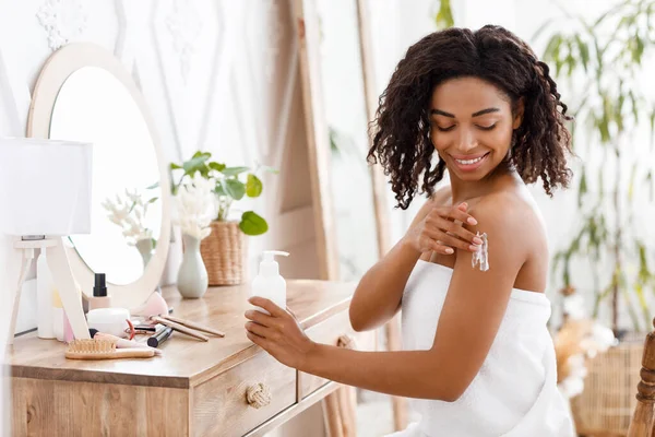 Διατροφή δέρματος. Χαμογελώντας ελκυστική μαύρη γυναίκα τυλιγμένο σε πετσέτα εφαρμογή λοσιόν σώματος — Φωτογραφία Αρχείου
