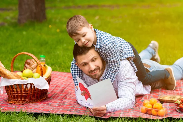 Grattis på fars dag. Söta barn kramar sin pappa, medan han läser gratulationskort på picknick i skogen — Stockfoto