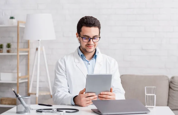 Консультация врача онлайн. Молодой человек в очках и белом халате с беспроводными наушниками смотрит на планшет — стоковое фото