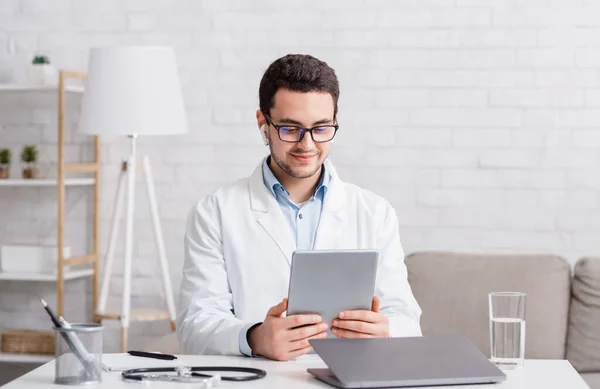 Consulta médica online. Jovem de óculos e casaco branco com fones de ouvido sem fio olha para tablet — Fotografia de Stock