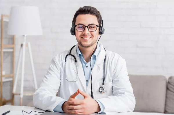Медицинская конференция или консультация онлайн. Молодой улыбающийся доктор в очках с наушниками — стоковое фото