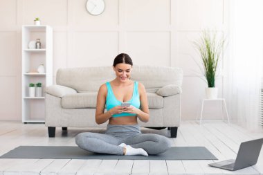 Evde oturan dizüstü bilgisayarda cep telefonu egzersizi yapan genç bir kadın.