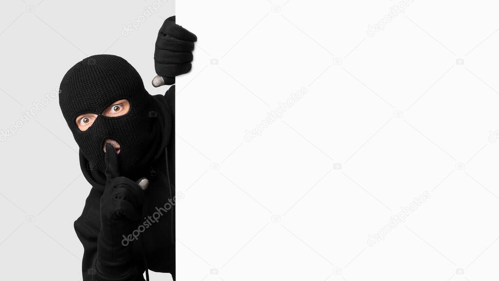 Masked thief peeking out white blank board, making hush sign