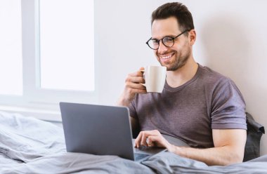 Laptopta çalışan, yatak odasında kahve içen neşeli adam.