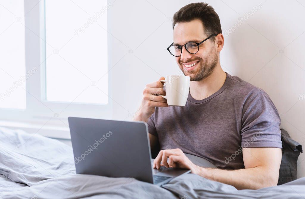 Cheerful Guy Working On Laptop Having Coffee Sitting In Bedroom