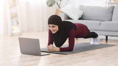 Ev Eğitimi. Fit müslüman kadın laptopun önünde yoga tahtası yapıyor.