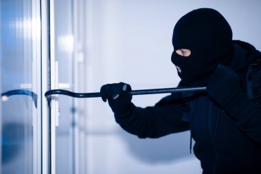 Siyah kar maskeli bir soyguncu levyeyle kapıyı kırıyor.