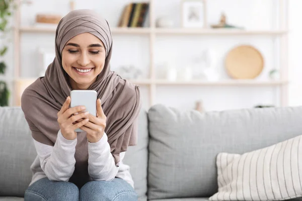 Веселая мусульманка в платке проводит время дома со смартфоном — стоковое фото