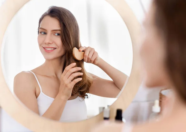 Vrolijke aantrekkelijke jonge vrouw kammen haar, kijken naar spiegel — Stockfoto