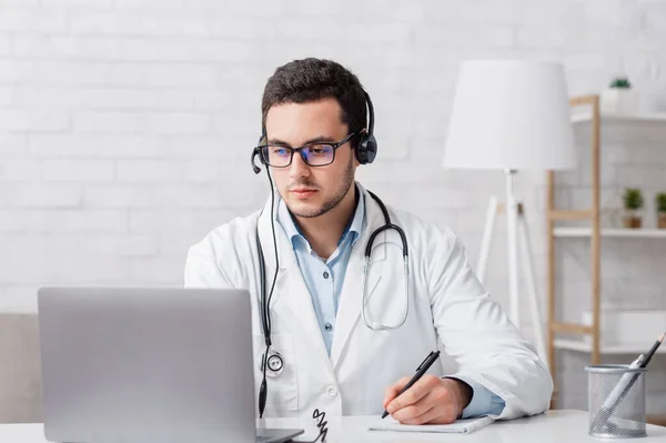 Patiëntenklachten online. Dokter maakt notities en kijkt naar laptop in het interieur van huis — Stockfoto