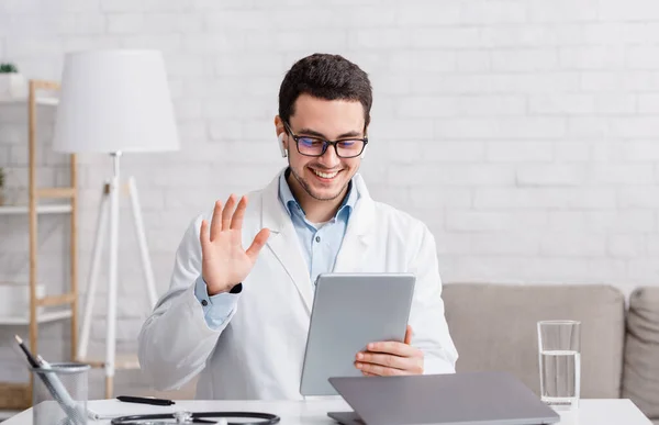 Tecnologias modernas e consulta. Médico sorrindo acena sua mão no tablet, respondendo a uma chamada de vídeo — Fotografia de Stock