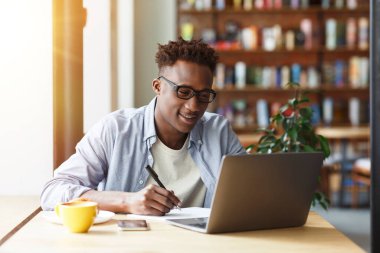 Milenyum Afrikalı Amerikalı öğrenci laptopuyla kafede kahvaltı yaparken not alıyor.