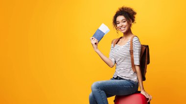 Seyahat çantasında oturan kadın biletleri sarı arka planda.