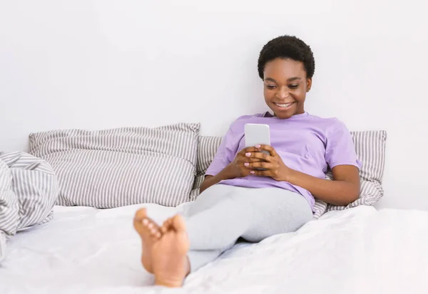 Navegar en internet en casa. Chica está acostado en la cama y el uso de teléfono inteligente —  Fotos de Stock