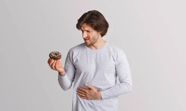 Man holds sweet donut in his hands and looks at it with pain, clutching his hands to stomach — Stock Photo, Image
