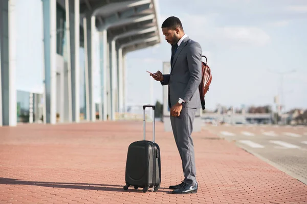 Gentleman africain utilisant un smartphone debout avec une valise de voyage à l'aéroport — Photo