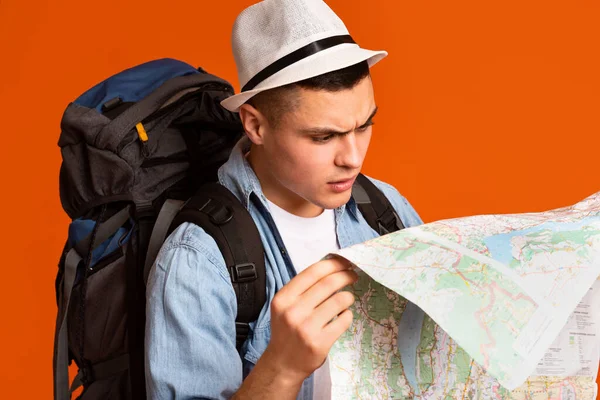 Nahaufnahme eines verwirrten Backpacker, der auf Karte über orangefarbenem Studiohintergrund blickt — Stockbild Verwirrter Backpacker schaut auf Karte über orangefarbenem Hintergrund — Stockfoto