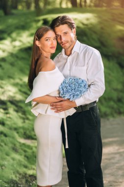 Yeni evli çift parkın dışında kameraya bakarak sarılıyor.