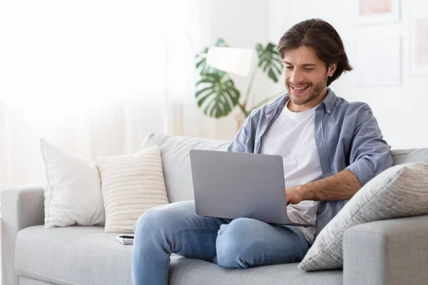 Hombre feliz escribiendo en el ordenador portátil, sentado en el sofá en casa — Foto de Stock