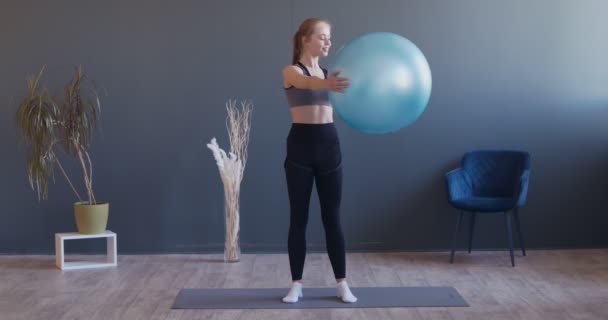 Молодая спортивная женщина занимается упражнениями, делая приседания с фитболом на руках в тренировочном зале — стоковое видео