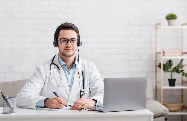 Online communication with patients. Doctor sits at table with laptop and takes notes — Stock Photo, Image