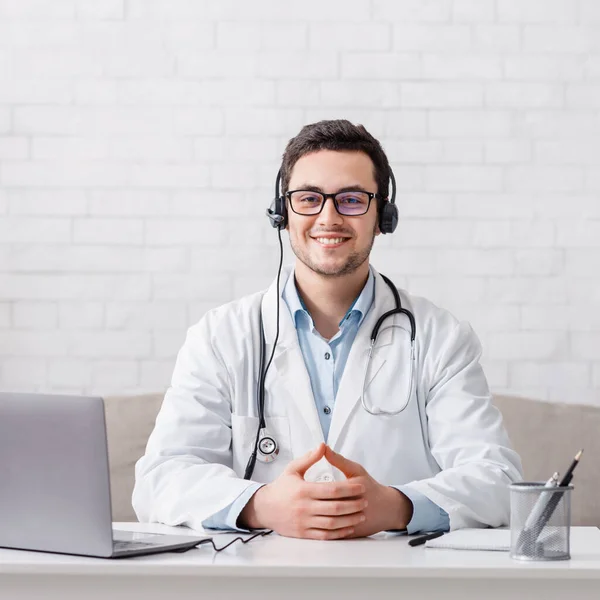 Blogg och svara på patientfrågor. Läkare i glasögon och hörlurar sitter vid bordet med laptop — Stockfoto