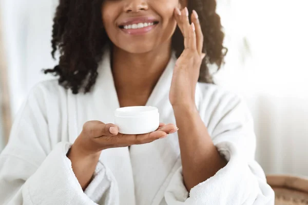 Donna nera che tiene il vaso di crema per la pelle biologica, applicando idratante sul viso . — Foto Stock
