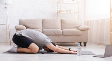 İnternetten ders alarak evde esnemek. Egzersiz yapan adam, yakınlarda dizüstü bilgisayar ve su.