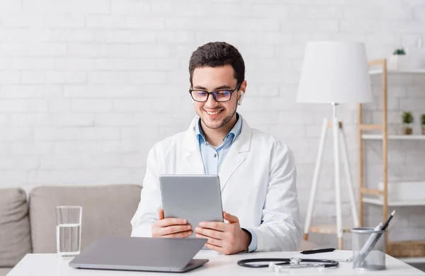 Online bezoek aan de dokter. Man kijkt naar tablet, zit aan tafel met laptop — Stockfoto