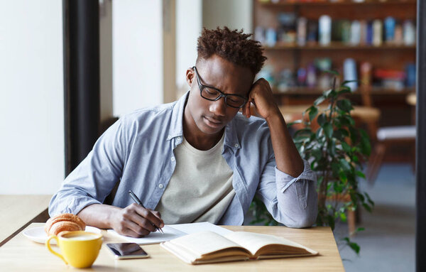 Focused African American student taking notes from book in cafe