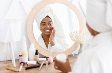 Young african woman applying moisturizer to her skin in front of mirror clipart