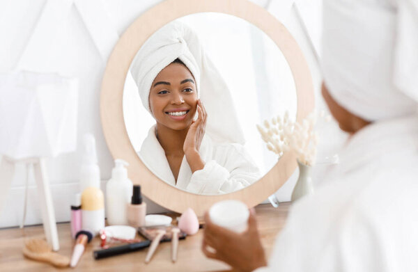 Young african woman applying moisturizer to her skin in front of mirror