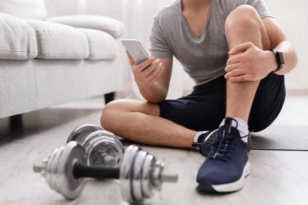 Trening mocy. Gość z nadajnikiem fitness i smartfonem siedzi na podłodze z hantlami — Zdjęcie stockowe