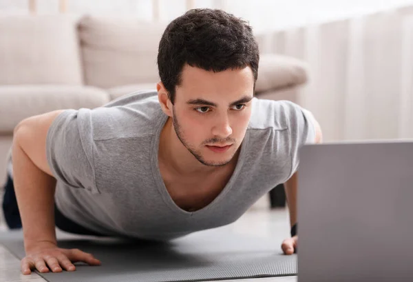 Trabajo remoto y deportes en línea. Chico es flexiones y mirando a la computadora portátil en el interior — Foto de Stock