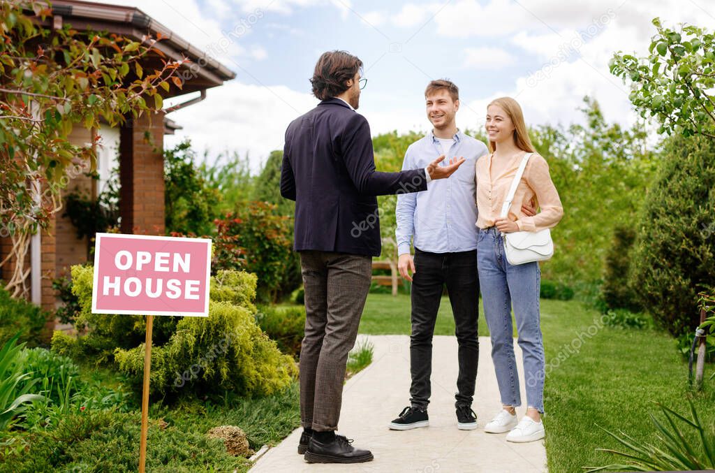 Confident realtor showing house for sale to newlywed young couple, outdoors