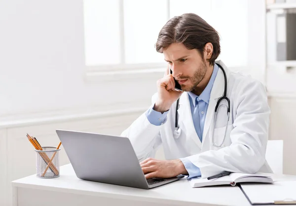 Focused doctor looking on results on laptop and talking on phone — Stock Photo, Image