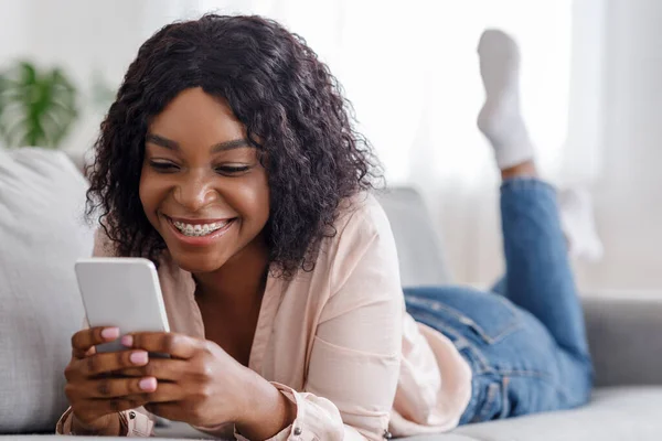 Menina preta feliz navegar na internet no smartphone enquanto deitado no sofá em casa — Fotografia de Stock