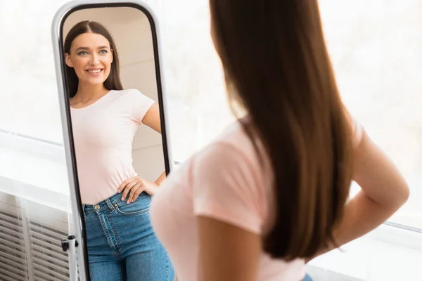 Улыбающаяся девушка смотрит в зеркало после того, как похудела стоя в помещении — стоковое фото